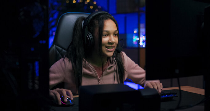 A teenage girl in a pink hoodie plays video games on her computer at night. She focuses on the screen, presses keys, and chats with online friends through a headset in a purple-lit room. - Powered by Adobe
