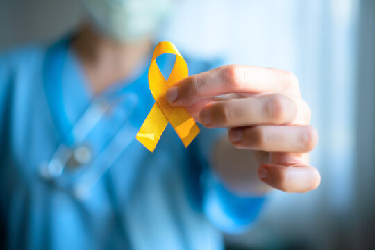 A nurse or doctor in medical attire holds a yellow ribbon representing bone diseases, childhood cancer, suicide prevention, and liver disease awareness.