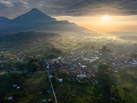 Aerial view of a village nestled amidst verdant rice terraces and shadowed forests, with a majestic mountain rising in the distance under a sky ablaze with dawn's golden light., East Java, Indonesia.