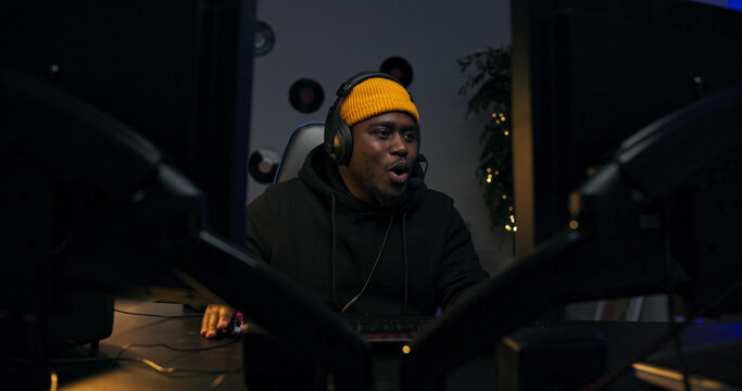 Focused gamer in cap and headphones talks to a friend while playing online. The dark room is lit by LED strips, and he flashes a bright smile while looking into the camera.