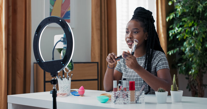 Makeup enthusiast creates video with ring light and phone. Girl highlights favorite brushes for foundation and bronzer, showing followers soft bristles in close-up view.