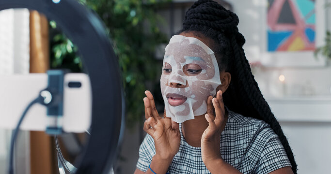 A beautiful young woman enjoys a beauty treatment at home, applying a sheet mask to her face while recording a skincare tutorial for her social media followers.