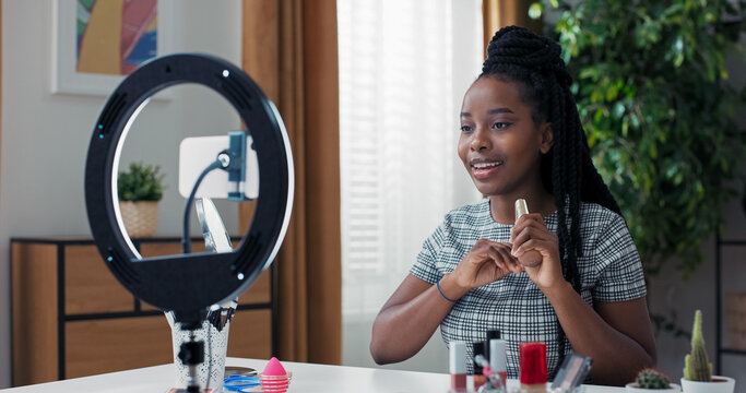 African American influencer films video promoting foundation for face, sharing brand recommendation, using cosmetics desk setup with bright ring light and smartphone tripod.