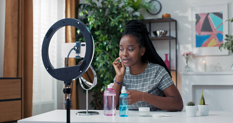 The female influencer shares a tutorial on properly removing foundation using cotton balls and makeup remover, recording a live video vlog with a phone tripod and ring light at home.