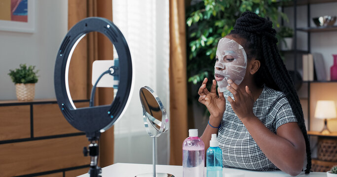 The beauty blogger introduces a facial mask to her audience. She smiles while recording content on social media with a ring light and smartphone.