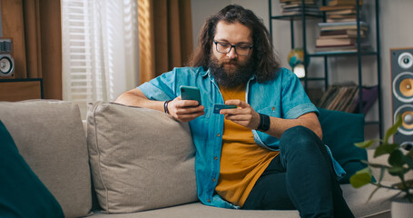 A man uses his smartphone to shop for clothes online, completing his purchase with a card while his dog joyfully jumps onto the couch to play.
