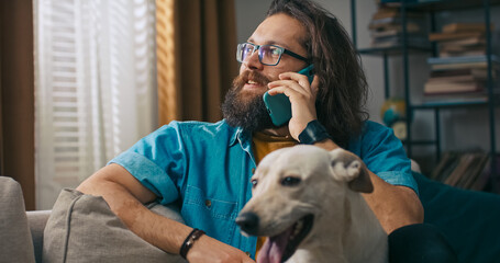A middle-aged man lounges at home and answers a call while his dog watches him, eager to go outside.