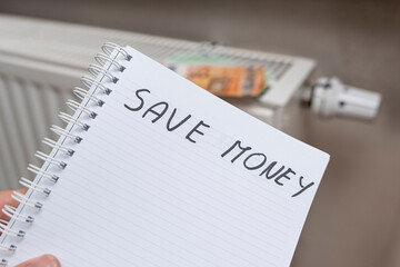 A handwritten note stating SAVE MONEY on a spiral notebook with a heating radiator in the background, highlighting a motivating financial message and practical savings tips.