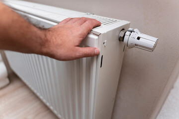 A hand adjusting the thermostat of a white radiator signifies the effort to regulate indoor heating. The scene takes place in a cozy room, reflecting home comfort.