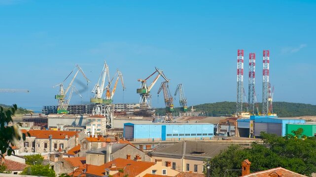 Shipyard cranes working in pula harbor. Tower cranes operating in the historic uljanik shipyard in pula, croatia, with the old town's rooftops in the foreground and a clear blue sky