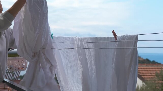Woman hanging freshly washed laundry on clothesline. Housewife hanging clean white laundry on a clothesline on the balcony of her house with a beautiful seaside view