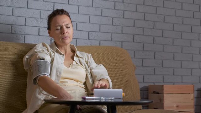 Woman measuring blood pressure feeling heart pain. Mature woman with a tonometer cuff on her arm sitting on the couch, holding her hand on her chest due to heart pain from high blood pressure