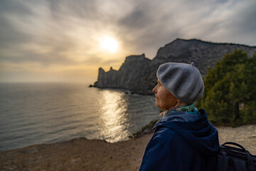 Elderly woman watches dramatic sunset over the seaside cliff looking contemplative and hopeful