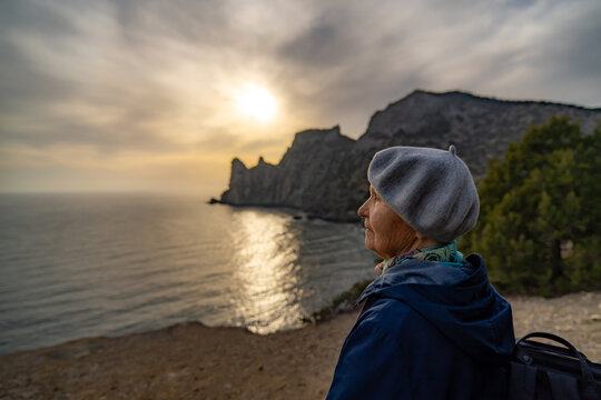 Elderly woman watches dramatic sunset over the seaside cliff looking contemplative and hopeful