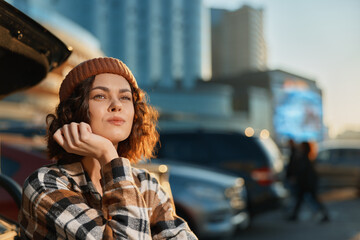 Woman portrait candid in urban street by open car trunk, beanie and plaid jacket, thoughtful gaze at golden hour glow, authenticity and mindful living, emotional storytelling lifestyle scene.