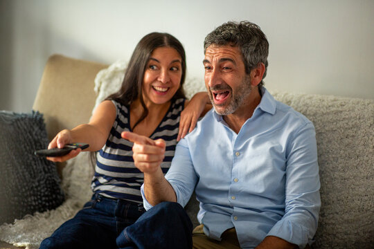 Happy couple laughing and pointing at television while watching together on sofa