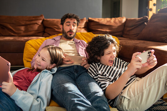 Adult father and children playing mobile games in living room, excited - Powered by Adobe