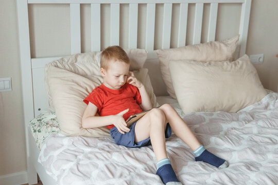 Young caucasian boy playing with a digital tablet while sitting on a comfortable bed during his leisure time, concept of childhood and technology.