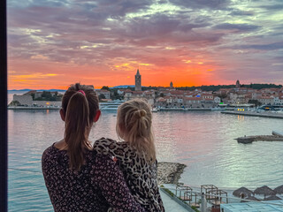Obraz premium A mother holds her young daughter while standing on a balcony to watch the vibrant red sunset over the historic skyline of Rab Old Town during a summer family vacation in coastal region of Croatia.