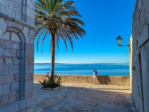 A mother holds her baby while standing near a large palm tree framed by ancient stone arches in Rab town. They look out at the calm blue Adriatic Sea under a clear sunny sky during a summer holiday.
