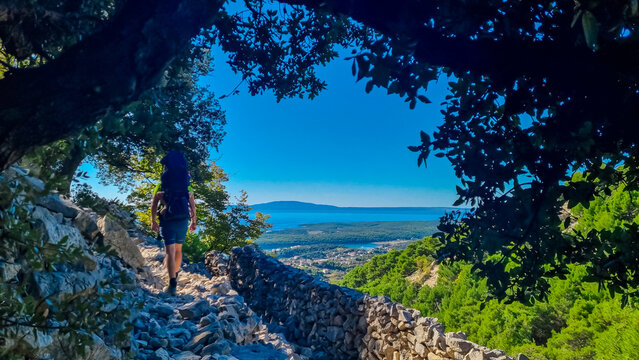 A hiker with a backpack treks along a rocky trail framed by green trees, heading towards a panoramic view of the blue Adriatic Sea and coastline on Rab Island, Croatia, on a sunny summer day.