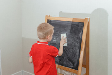 Blond boy erasing letter A from a chalkboard with an eraser. Early education, learning for child. Elementary school concept for student.