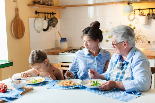Happy multigenerational family eating dinner together at home kitchen. Three generations enjoying pasta, healthy food. Woman, child and grandmother have fun. Candid, authentic lifestyle moment