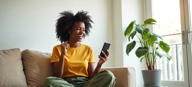 Young african american woman laughs on sofa holding smartphone. Celebrates good news, win, success. Female enjoys online content, watching video, playing mobile game. Excited reaction, positive
