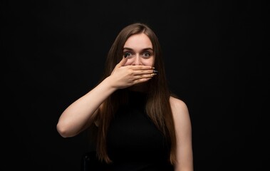 Surprised young woman covering mouth with hand in shocked emotional reaction on dark studio background