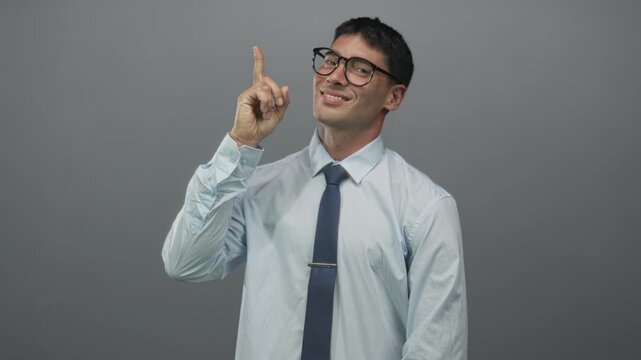 Man points finger upward in grey studio while wearing glasses and tie and smiling at ceiling; optimism.