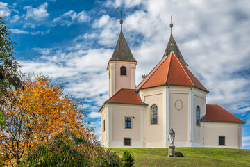Baroque church with red roof and autumn trees under blue sky
