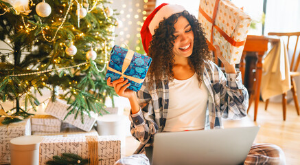 Portrait of woman in pajamas and Santa hat with laptop surrounded by holiday gifts in cozy room. Happy woman works on laptop near decorated Christmas tree. Holiday concept, freelance.