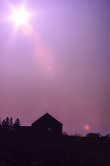 Starburst sun in a purple and pink sky with lens flare above a silhouetted barn.