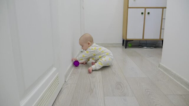 Active baby boy sits on floor, happily playing with a purple ball. Looking at camera and funny smiling. Developing motor skills through tactile exploration and play. Safe home environment for infant