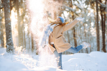 A cheerful woman in warm clothing is having fun with snow in a sunny forest. Beautiful woman enjoys...