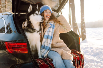 A happy woman and her pet sit on a blanket in the trunk of a car. A young woman in warm clothes with her husky enjoys a snowy day in the forest on a road trip. Concept of friendship, fun, adventure. © maxbelchenko