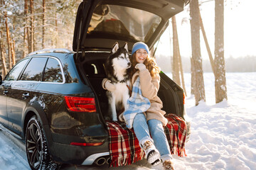 A happy woman and her pet sit on a blanket in the trunk of a car. A young woman in warm clothes...