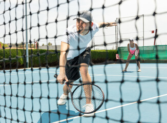 Portrait of emotional determined young guy playing tennis on open court in summer, swinging racket to return ball over net. Sportsman ready to hit volley.