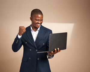 A young Nigerian man in a navy pinstriped suit stands in a studio holding a closed laptop at his side while facing the camera under soft directional lighting.
