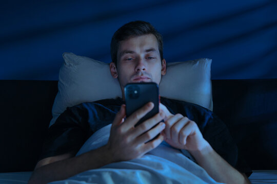 A tired man lies awake in a dark bedroom, staring at his glowing smartphone and doom scrolling late at night despite his growing fatigue. Digital addiction or insomnia concept. Copy space 