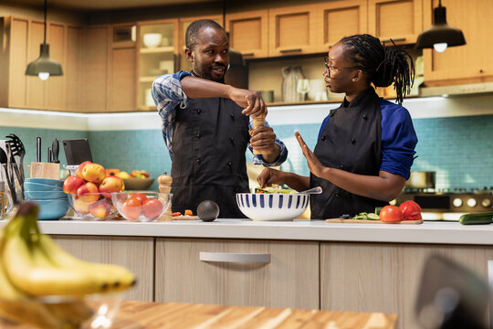 African american man adding pepper to the salad and woman stopping him, enough seasoning for healthy lunch meal in bowl. Young jolly couple preparing food together, wearing aprons. - Powered by Adobe