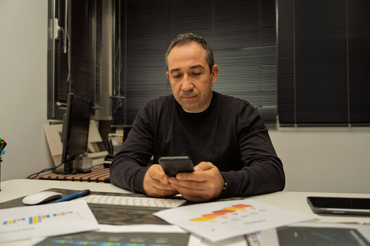 Mature man working, browsing smartphone at office desk