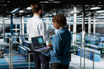 African american IT professional working on pc in high tech data center, handling maintenance tasks to ensure optimal performance for server automation. Female checking code on laptop.
