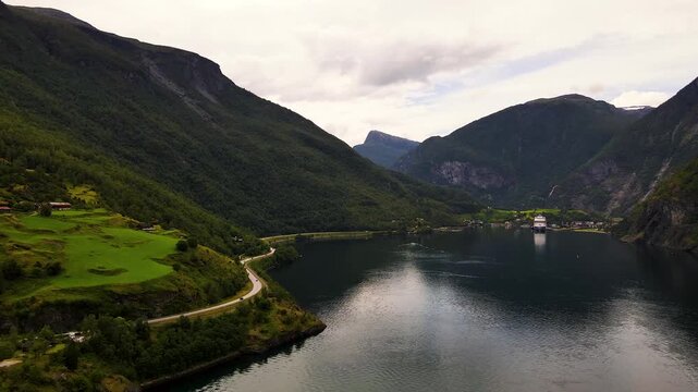 Aerial view of a cruise ship docked on the still waters of a fjord surrounded by lush green mountains and a winding road in Flam, Vestland, Norway.