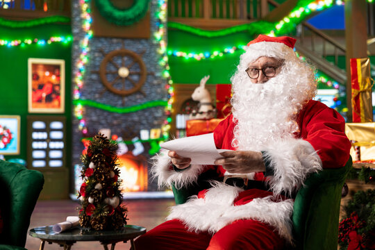 Portrait of senescent man playing Santa Claus role opening letter from kids asking for presents. Senior performer wearing Santa costume in Christmas ornate room reading gift wish lists from children
