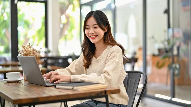 カフェの明るい窓際でベージュのセーターを着た若い女性がノートパソコンに集中して入力している