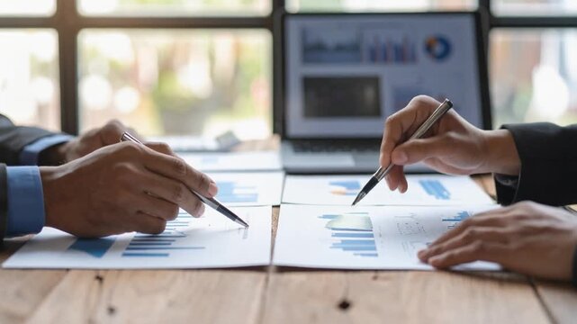 木製のテーブルに広げられた資料を熱心に確認する二人の男性の腕と手元 奥にはノートパソコンの画面が見えるビジネスミーティングの様子