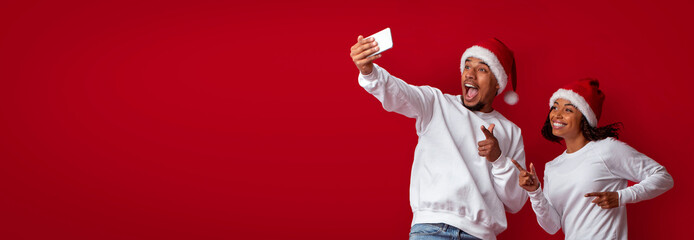 Two friends take a cheerful selfie against a bright red background, both wearing Santa hats and...