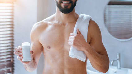 A man stands in a modern bathroom, holding a stick of deodorant in one hand and a white towel over...