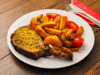 Fine dinner with spicy pork chop, potato wedges and fresh tomato on a plate on a wooden table. Tasty meal with meat and vegetables.